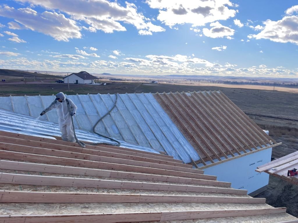 Spray-foam crew member in PPE applying closed-cell foam to the topside roof deck of a Moorcroft, Wyoming home with open prairie in the distance.