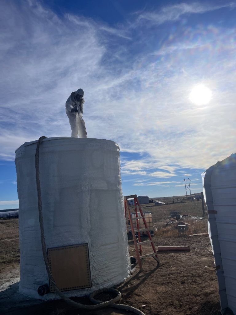 Seamless Systems crew member in full PPE spraying closed-cell foam onto the top of a curved metal oil storage tank on a Wyoming prairie site, sun flaring over the horizon.